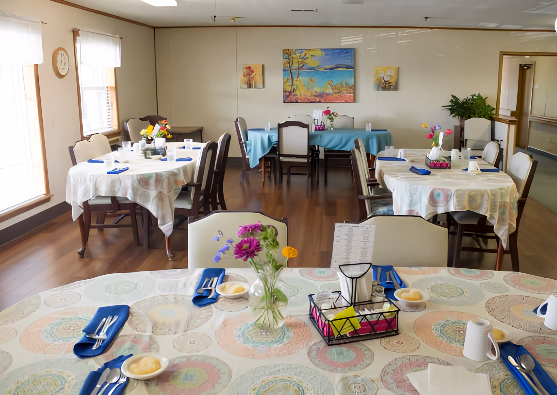 A dining room with several round tables covered with patterned tablecloths and set with blue napkins, silverware, and small bowls of applesauce. Each table has a small vase with colorful flowers. The room has wooden floors, light-colored walls, and windows with white curtains. There are paintings on the walls and a green plant in the corner.