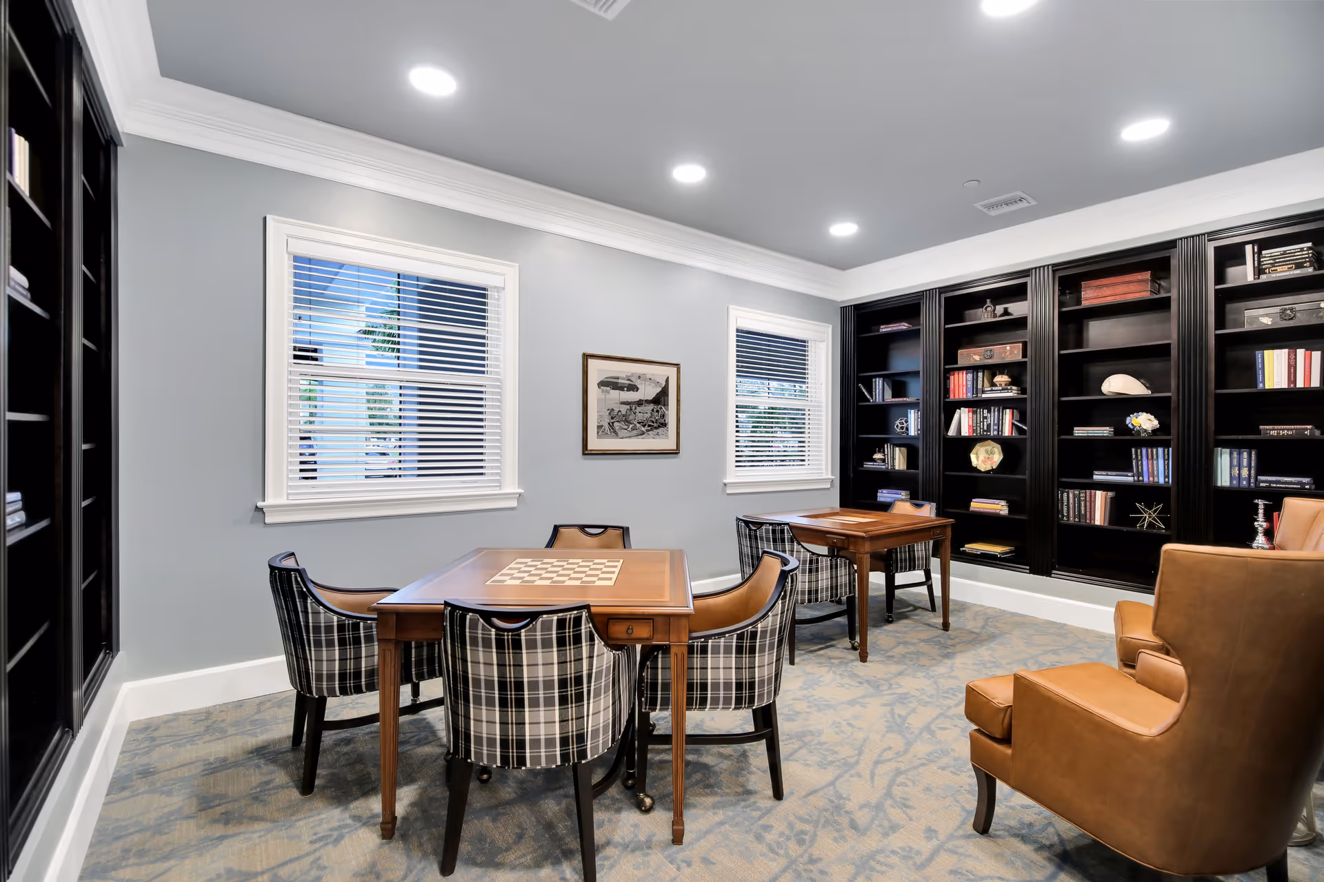 A cozy interior room with two wooden tables featuring built-in chessboards, surrounded by plaid upholstered chairs. The room has light gray walls, two windows with white blinds, and built-in black bookshelves filled with books and decorative items. There is also a brown leather armchair in the foreground.