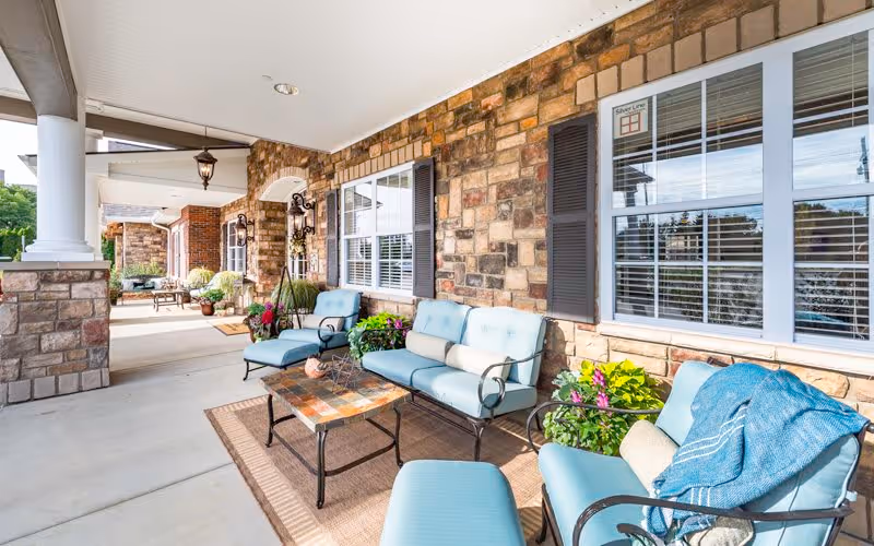 Covered outdoor patio area with stone walls and white columns, featuring cushioned blue seating including chairs and a loveseat around a small coffee table on a rug. There are potted plants and flowers decorating the space, with large windows and black shutters on the wall behind the seating.