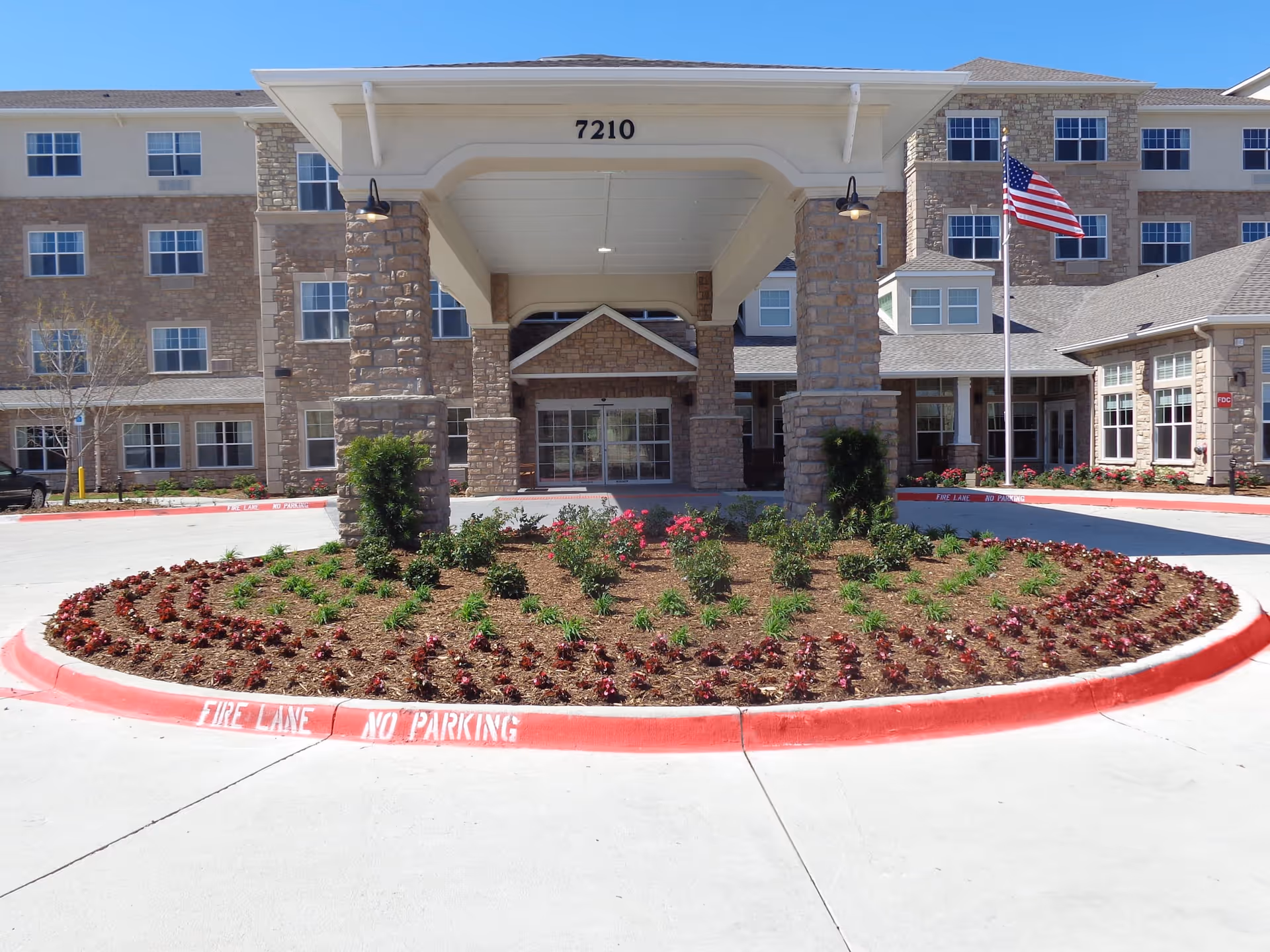 Front entrance of a multi-story senior living facility with stone pillars supporting a covered driveway. A landscaped circular flower bed with small plants and flowers is in the foreground. An American flag is flying on a flagpole to the right of the entrance. The building number 7210 is displayed above the entrance.
