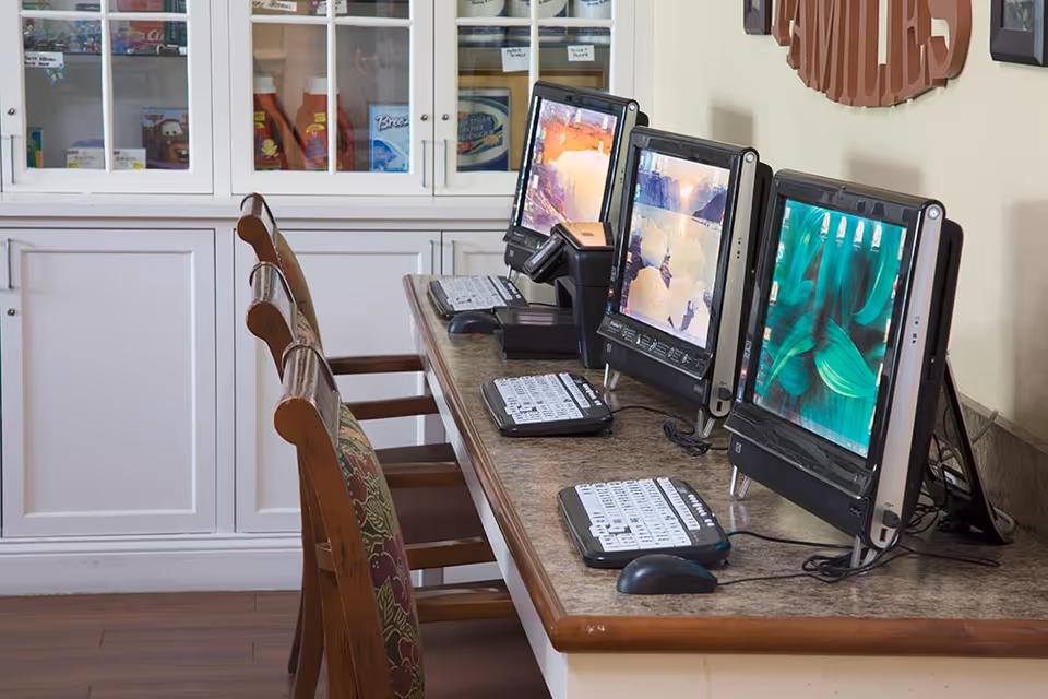 A row of three desktop computers with large screens and adaptive keyboards on a long table with wooden chairs. Behind the table is a white cabinet with glass doors displaying various items inside.