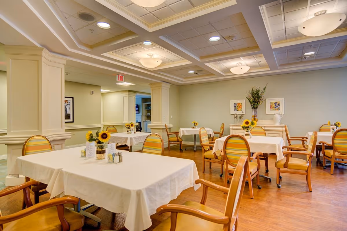 Bright dining room with tables covered in white tablecloths, wooden chairs, and sunflower centerpieces.