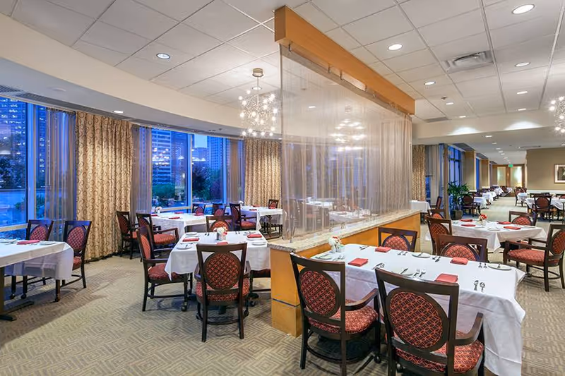 A spacious dining room with multiple tables covered in white tablecloths, each set with red napkins and silverware. The room features large windows with patterned curtains, modern chandeliers, and a transparent divider separating sections of the dining area. The setting appears elegant and well-lit, suitable for a senior living facility.