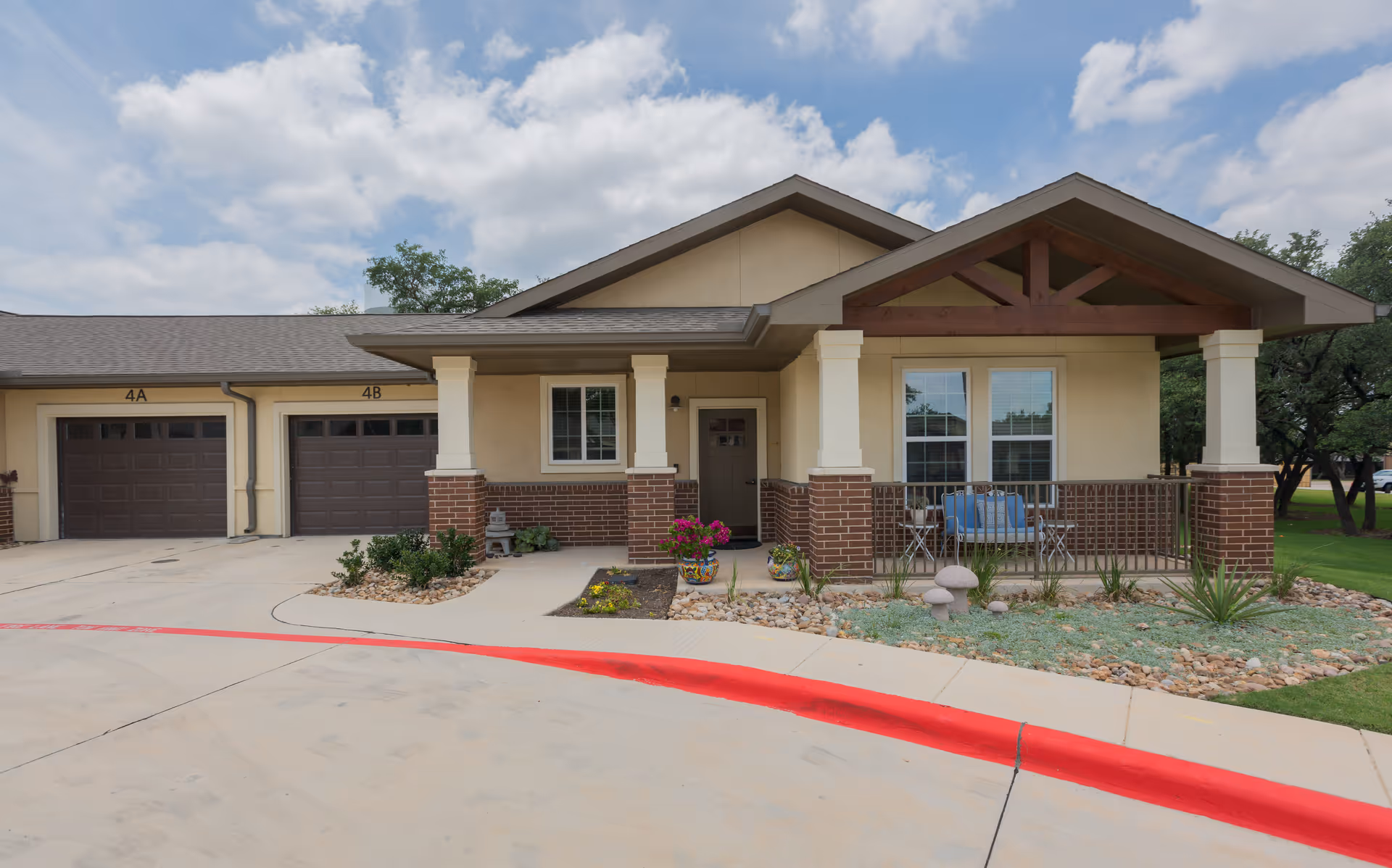 Exterior view of a single-story senior living facility building with a covered porch, brick and beige walls, two garage doors labeled 4A and 4B, a small garden with plants and flowers, and a curved driveway with a red painted curb under a partly cloudy sky.