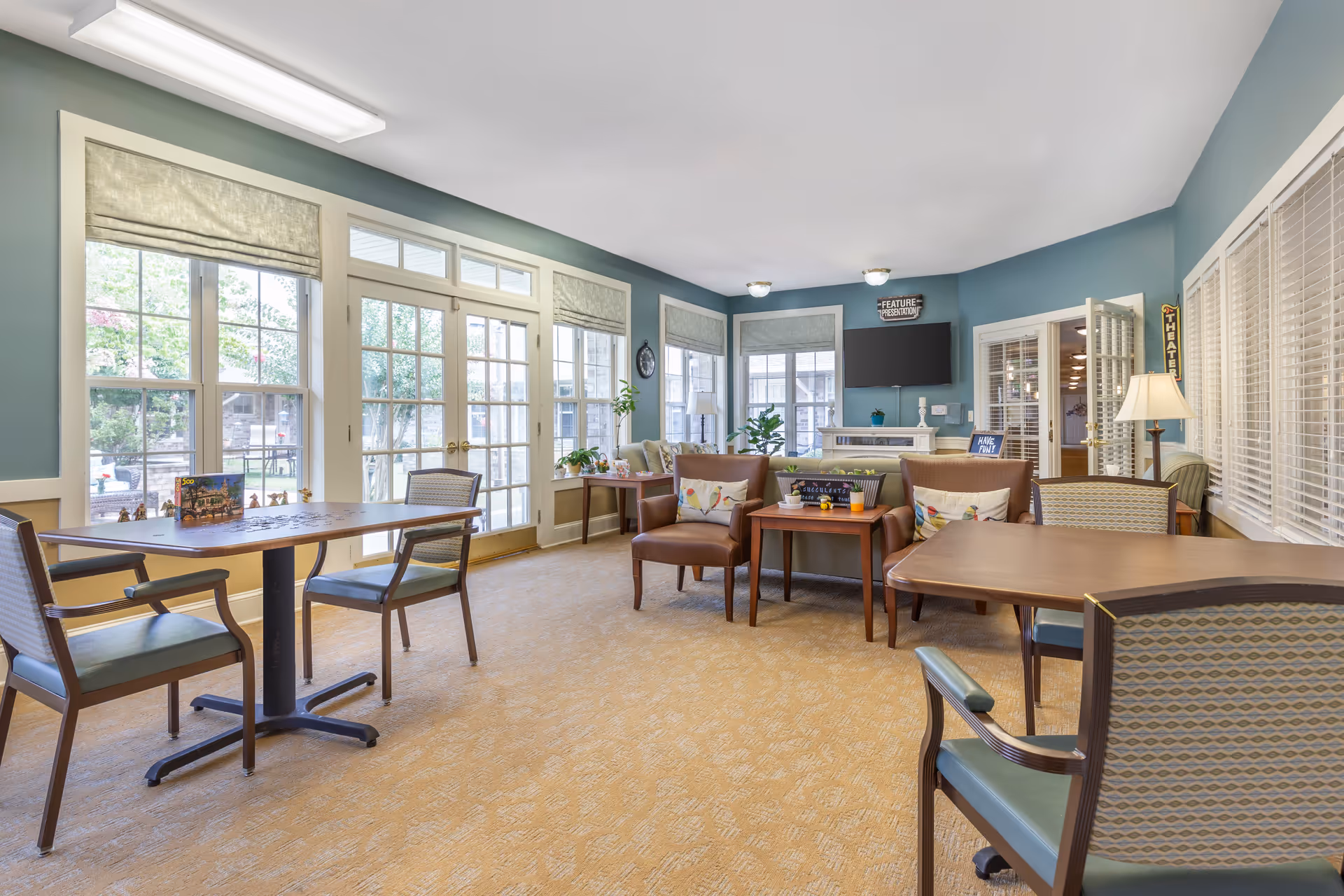 Bright senior living communal lounge with tables, chairs, armchairs, a TV, and large windows.