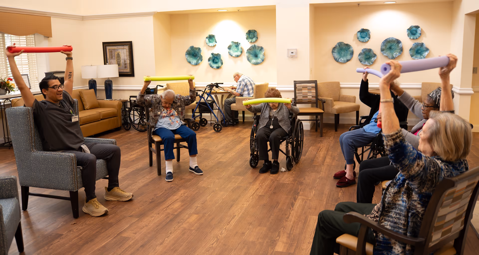 A group of elderly individuals and a caregiver participating in a seated exercise session in a senior living facility common room. They are holding colorful foam noodles above their heads while seated in chairs and wheelchairs. The room has wooden flooring, beige walls, and decorative blue wall art.