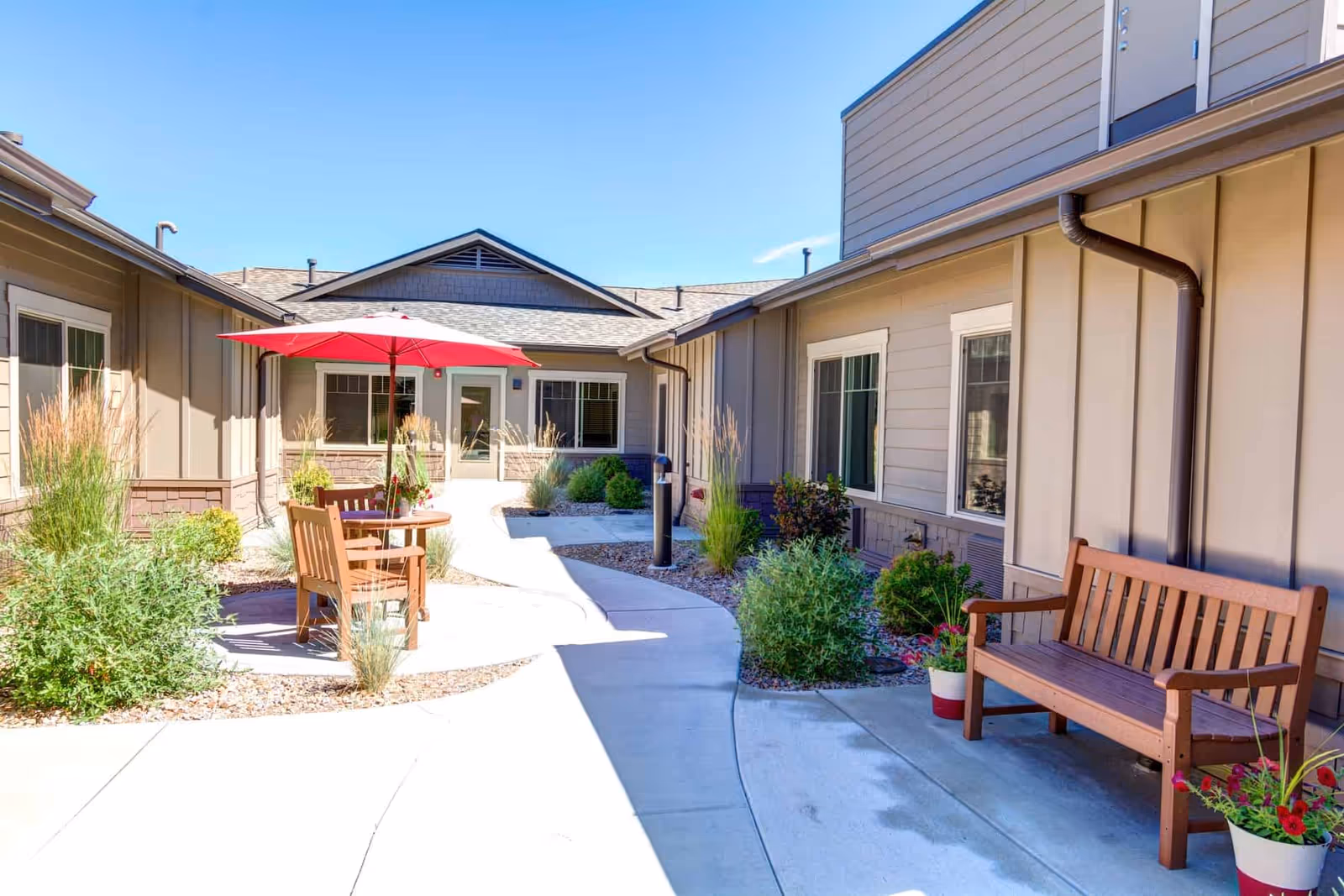 Sunny courtyard at an assisted living facility with benches, a table under a red umbrella, and landscaped walkways around the building.