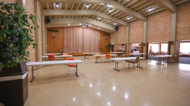 A large multipurpose room with a stage at the far end, featuring a closed curtain and an American flag. The room has a high ceiling with exposed beams and brick walls. Several long tables and chairs are spaced out across the polished floor. There are large windows and an open doorway leading to another room with more tables and chairs. A potted plant is visible on the left side of the image.