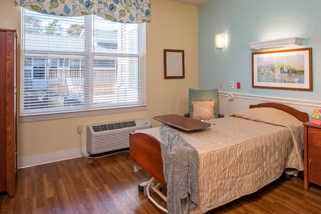 A well-lit bedroom in a senior living facility featuring a single bed with a beige quilt, a wooden headboard, and a gray knitted blanket draped over the foot. Next to the bed is a wooden nightstand with a colorful item on it. A green chair with a pink cushion is placed against the wall, which is painted light blue and beige. Above the bed hangs a framed painting of boats on water. A large window with white blinds and a floral valance lets in natural light, and a white air conditioning unit is installed below the window. The floor is wooden.
