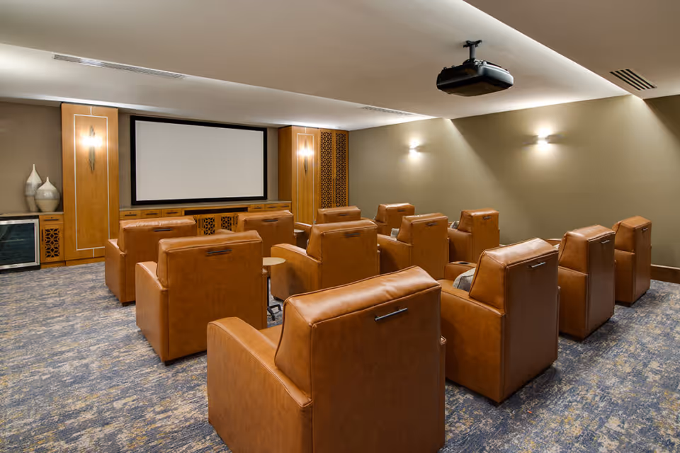 A small theater room with two rows of brown leather recliner chairs facing a large blank projector screen. The room has soft lighting on the walls, a ceiling-mounted projector, and wooden cabinetry with decorative vases on the left side.