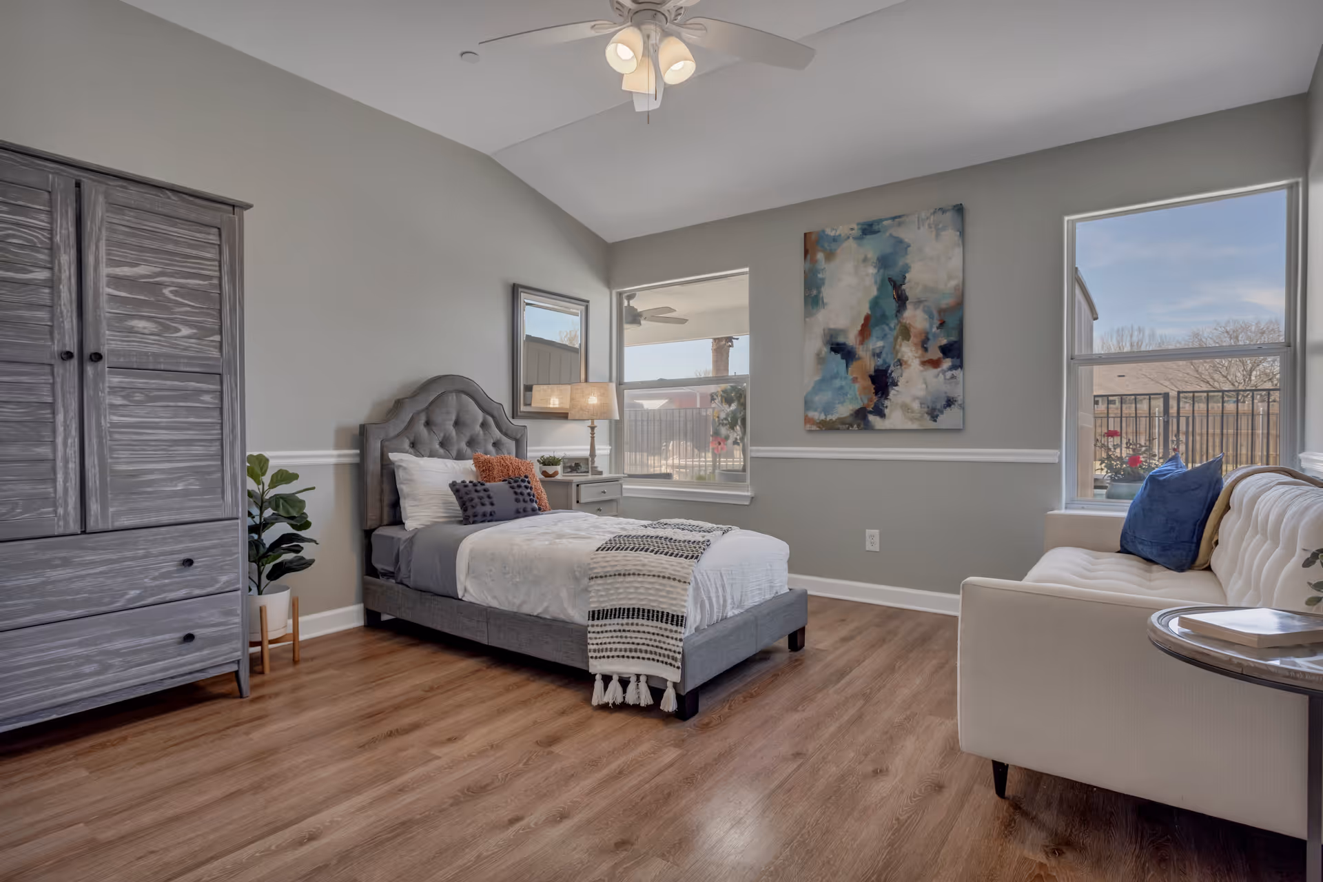 A bright and spacious bedroom with a gray upholstered bed, white bedding, and decorative pillows. There is a wooden armoire on the left, a small nightstand with a lamp and plant next to the bed, and a white tufted sofa with blue and beige pillows on the right. The room has light gray walls, two large windows letting in natural light, a ceiling fan with lights, and a colorful abstract painting on the wall.