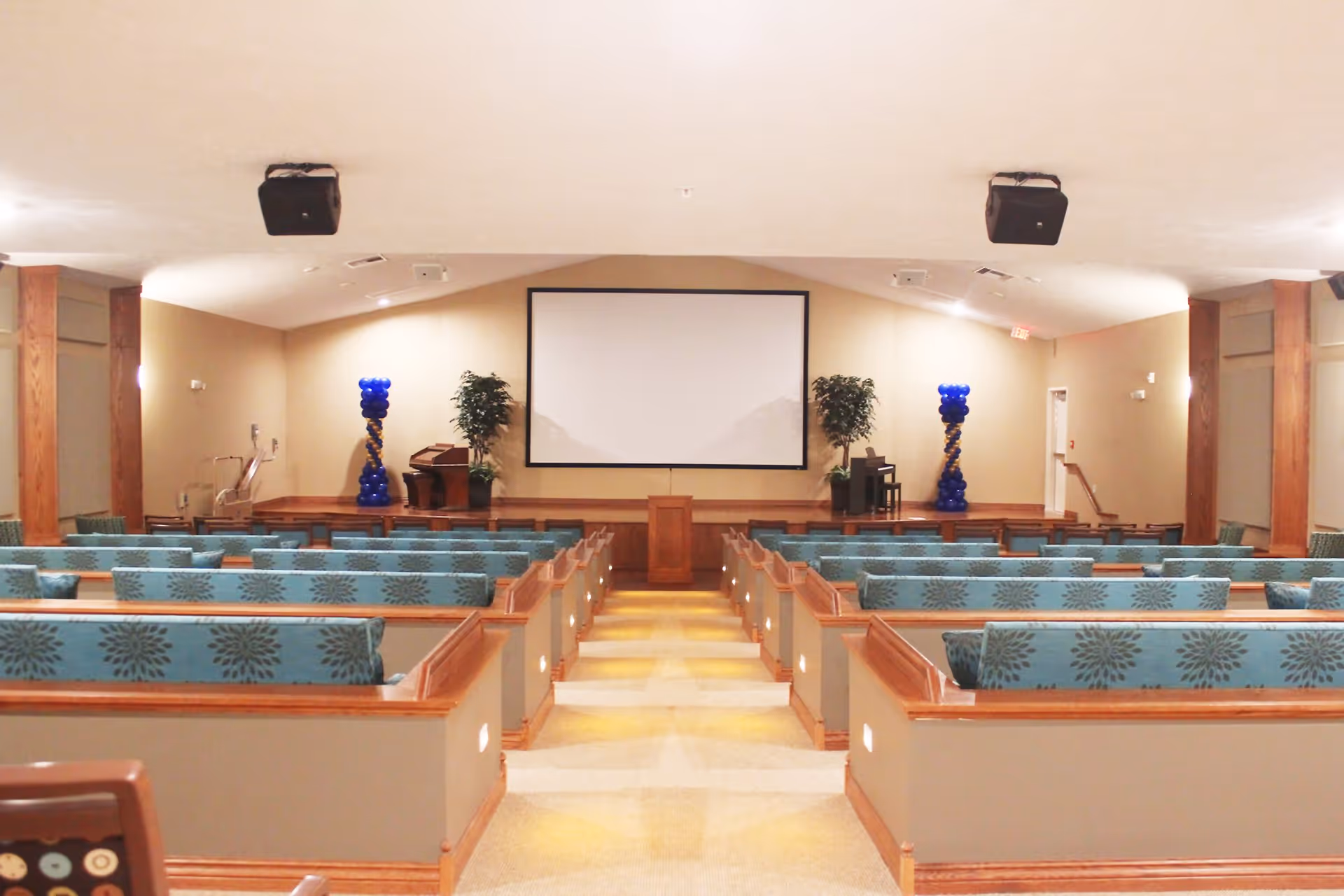 Interior view of a large meeting or presentation room with rows of cushioned benches facing a stage with a podium and a large projection screen. The room has beige walls, wooden accents, two potted plants, and blue balloon decorations on either side of the stage.