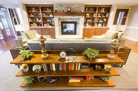 A cozy living room with a gray sofa facing a stone fireplace surrounded by built-in wooden bookshelves filled with books and decorative items. Behind the sofa is a wooden console table with plants, decorative objects, and books. The room has warm lighting and a carpeted floor.