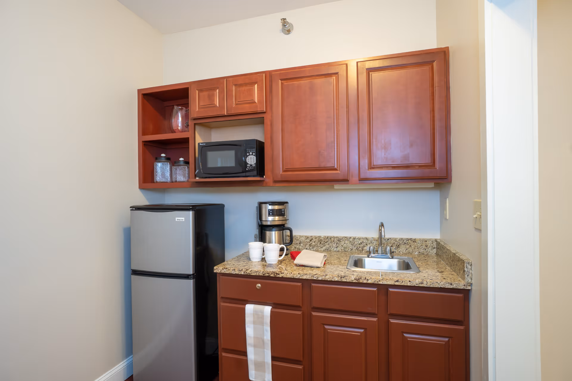 A small kitchenette with wooden cabinets, a granite countertop, a stainless steel mini refrigerator, a microwave, a coffee maker, two white mugs, and a sink. There is a white and beige striped towel hanging from one of the cabinet handles.