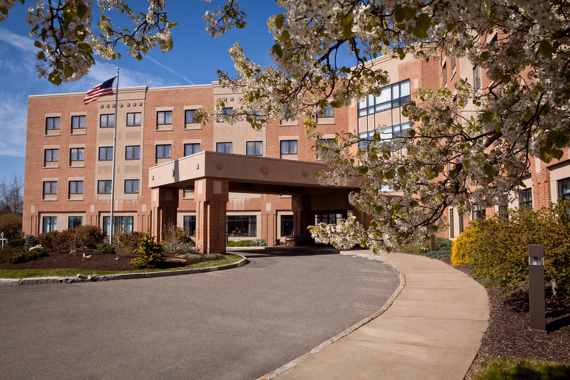 Exterior view of a multi-story brick building with a covered entrance and an American flag on a flagpole. The building is surrounded by landscaped bushes and trees with white blossoms, and a curved driveway and sidewalk lead to the entrance.