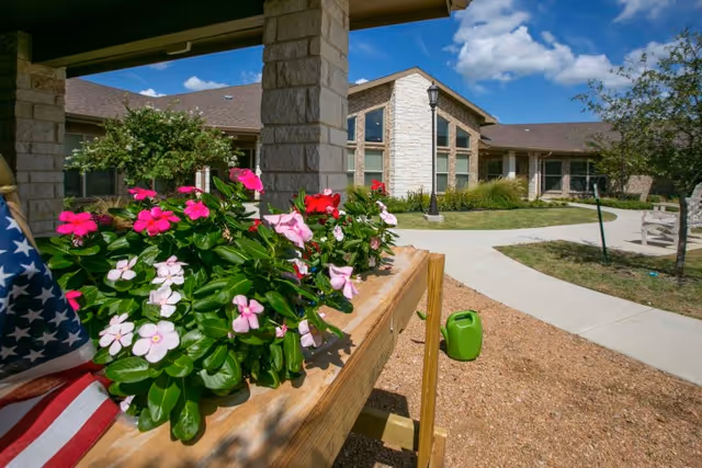 Stone-faced senior living building viewed from a sunny courtyard with a wooden planter of pink flowers in the foreground.