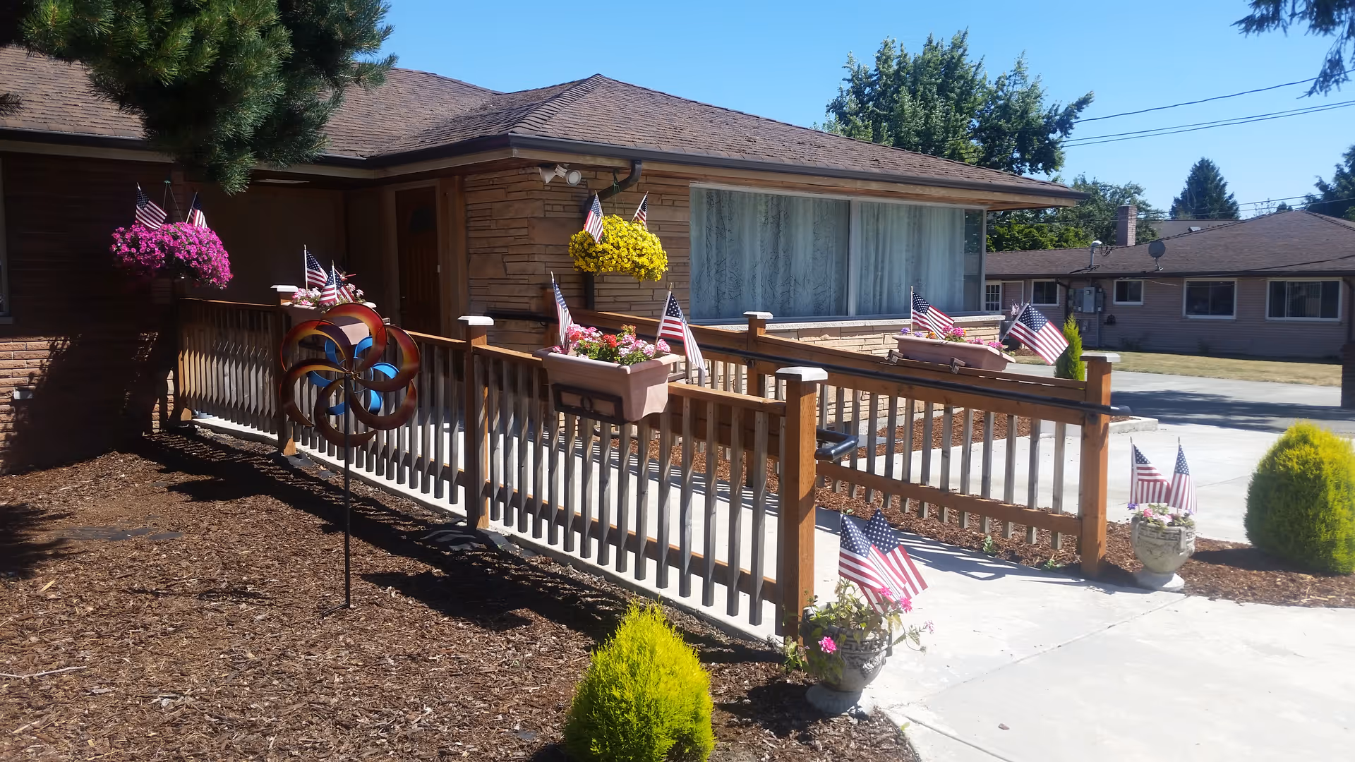 Front exterior of a single-story building with a wooden wheelchair ramp decorated with American flags and potted flowers on a sunny day.