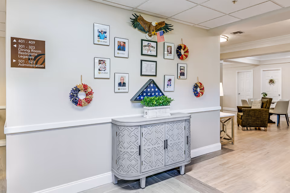 Interior hallway of a senior living facility with a decorative cabinet against the wall. Above the cabinet are framed photos of veterans, a folded American flag in a display case, patriotic wreaths, and an eagle decoration holding a small American flag. A sign on the wall indicates directions to rooms, dining room, restrooms, legacies, and administration. In the background, there is a seating area with chairs and tables.