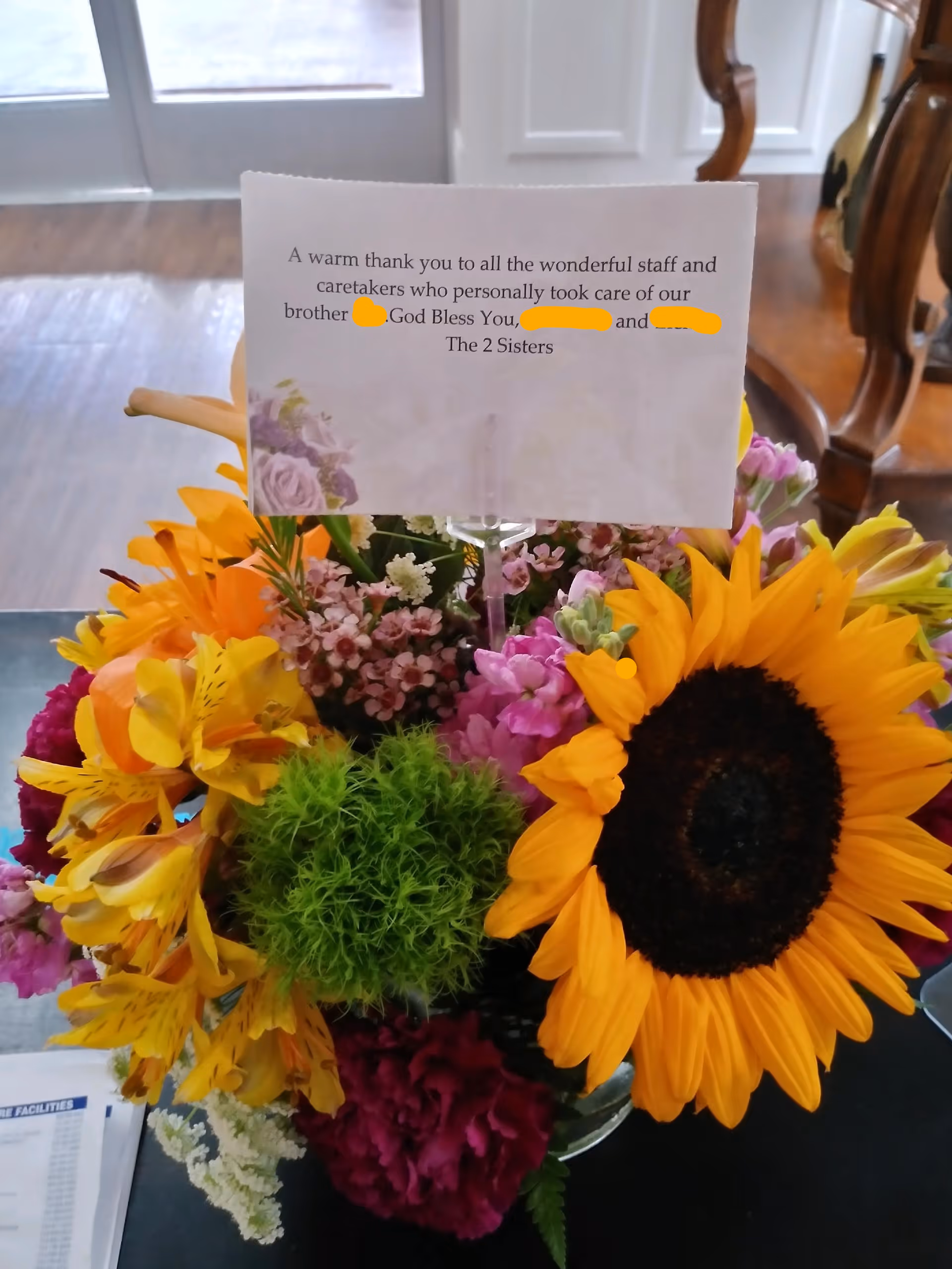 A colorful bouquet of flowers including a large sunflower, yellow lilies, pink and purple flowers, and green foliage in a glass vase on a table. A card is attached to the bouquet with a message thanking the staff and caretakers for their care. The background shows a wooden floor and part of a chair.