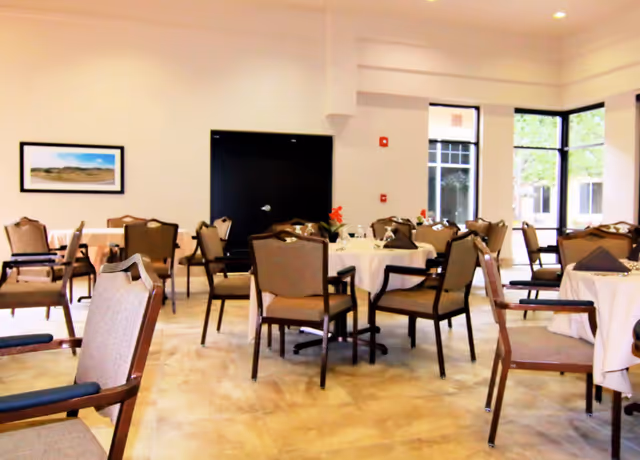 A bright dining room with several round tables covered with white tablecloths, each surrounded by brown cushioned chairs. The room has large windows letting in natural light and a framed landscape picture on the wall.