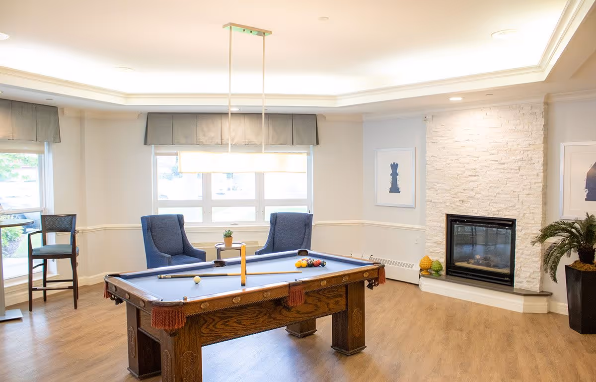 A bright and spacious room with a pool table in the center, two blue armchairs near a window, a modern fireplace with white stone surround, and a potted plant in the corner. The room has light-colored walls, wood flooring, and a rectangular ceiling light fixture above the pool table.