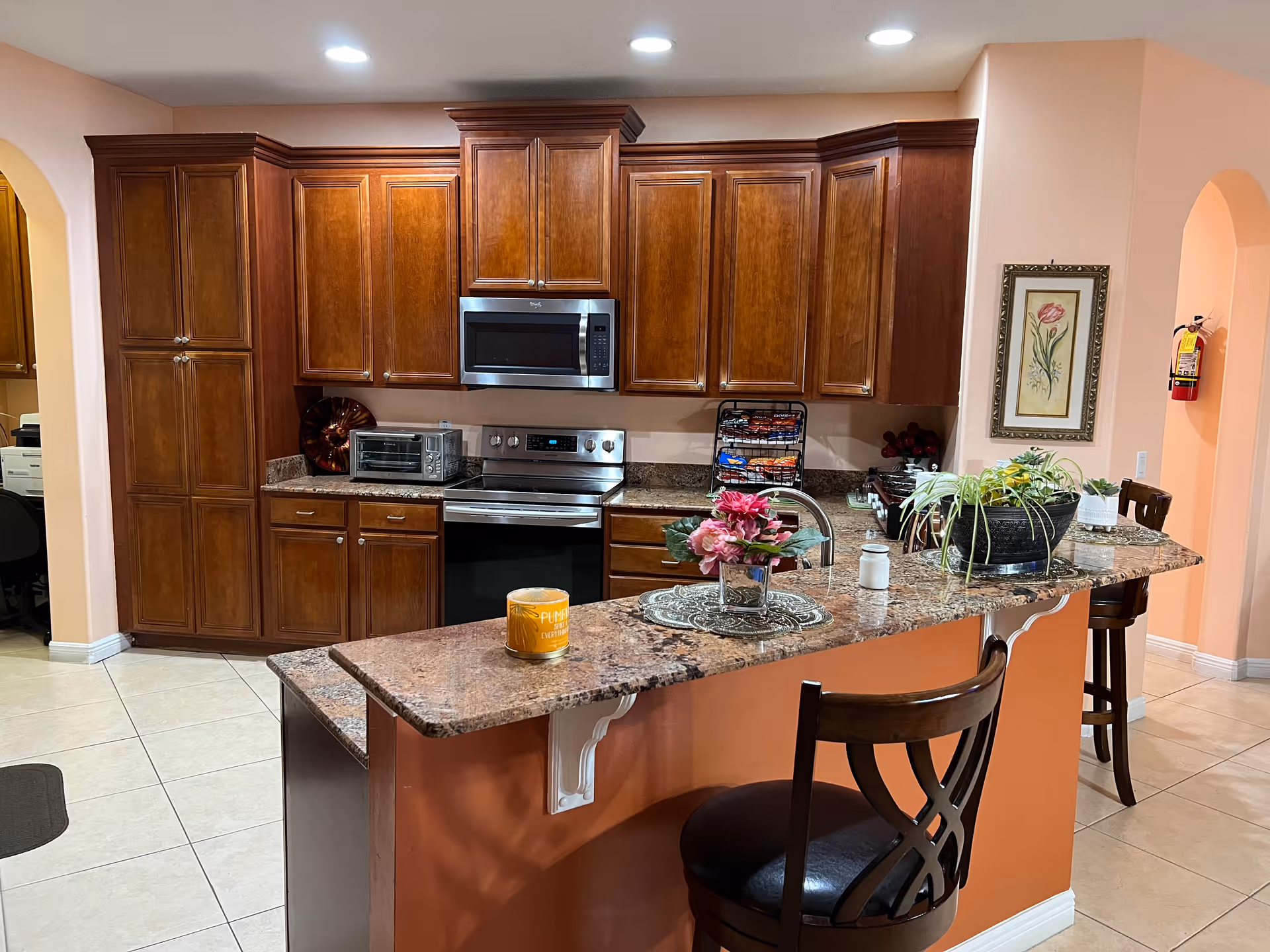 Kitchen with cherry wood cabinets, stainless steel appliances, and a granite-topped breakfast bar with two stools and decorative plants.