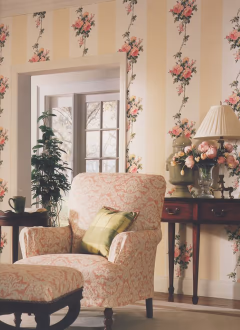 A cozy living room corner with a floral patterned armchair and matching ottoman. The armchair has a green plaid cushion. Behind the chair is a wooden side table with a green ceramic lamp, a vase of pink flowers, and a table lamp with a pleated shade. The walls are decorated with yellow and cream vertical stripes and floral wallpaper. A window with multiple panes and a potted plant are visible in the background.
