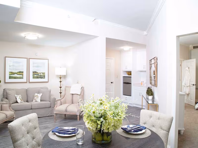 Light-filled living and dining area with a round table topped by a floral centerpiece, sofas and armchairs, and a small kitchenette in the background.