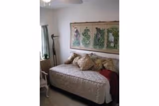 A small bedroom with a single bed covered in a beige quilt and multiple pillows. Above the bed is a framed artwork featuring botanical prints. A window with green curtains and a floor lamp are visible to the left of the bed.