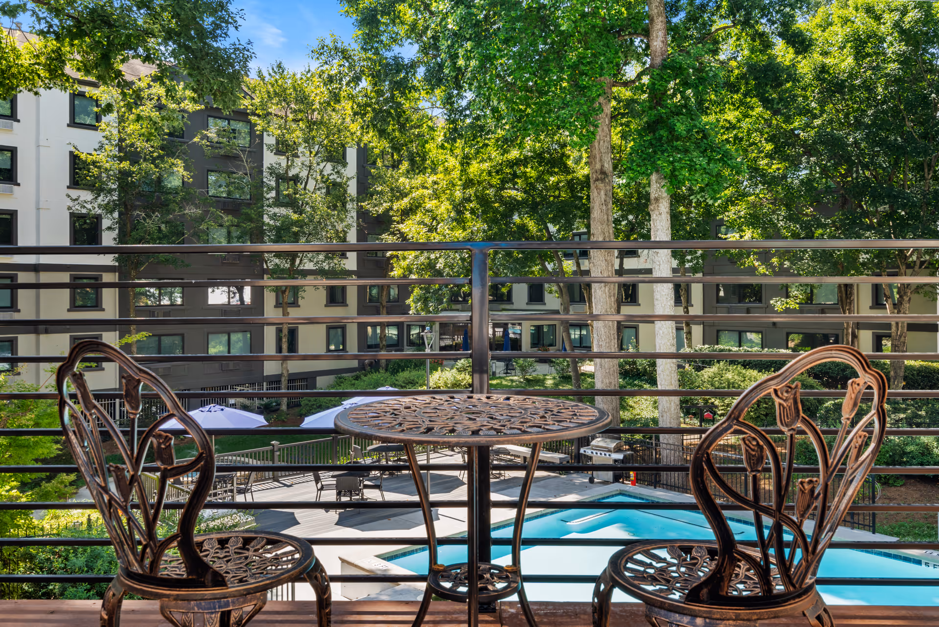 View from a balcony with two ornate metal chairs and a matching round table overlooking a swimming pool surrounded by trees and a multi-story residential building in the background.
