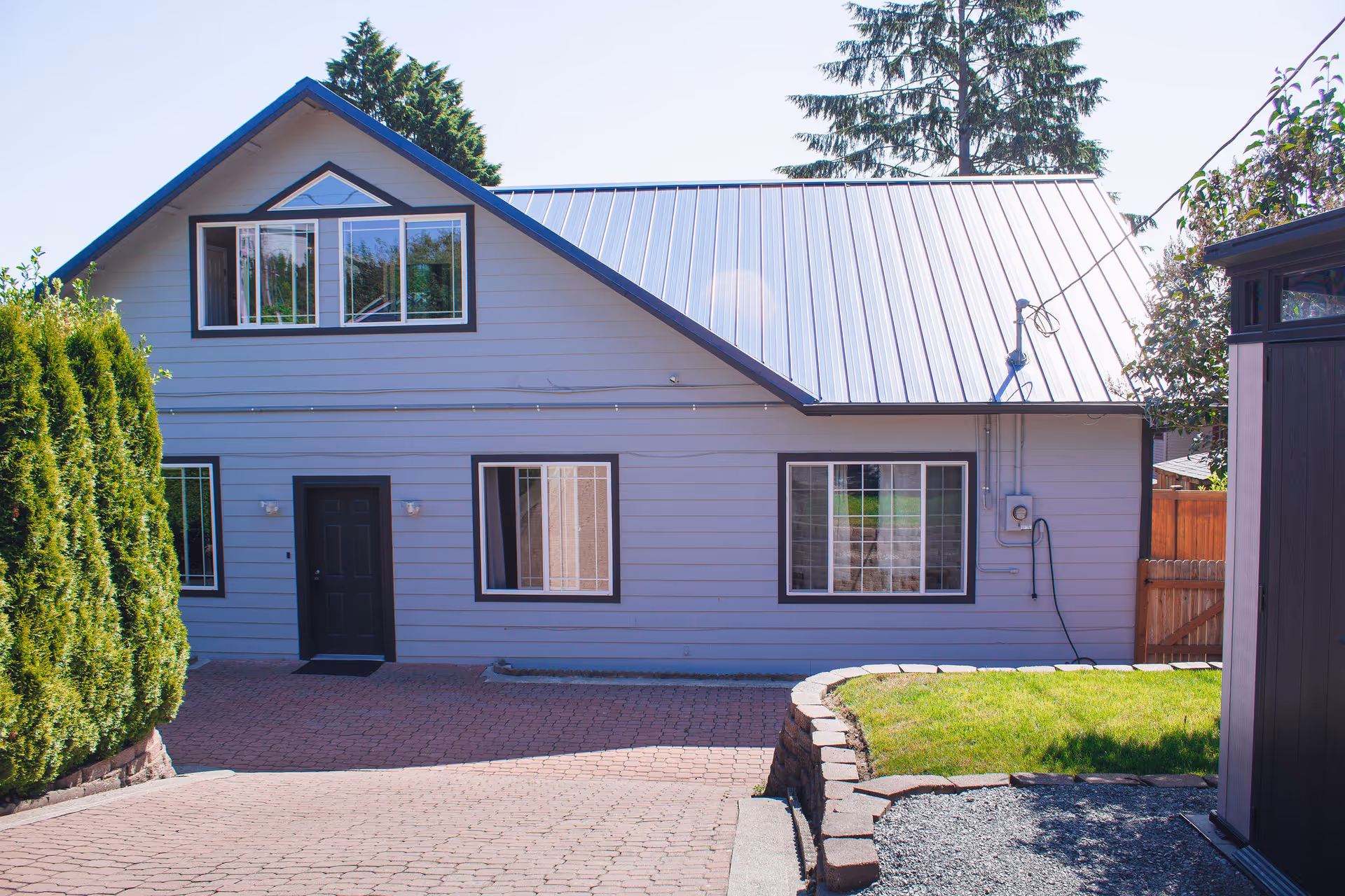 Front exterior of a light-gray two-story house with a metal roof, multiple windows, a black front door, and a paved driveway.