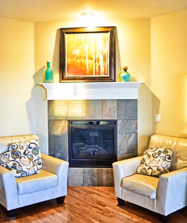 A cozy living room corner featuring two beige armchairs with patterned cushions placed on either side of a tiled fireplace. Above the fireplace mantel, there is a framed painting with warm tones and two decorative vases, one green and one blue, on each side of the mantel. The floor is wooden and the walls are painted a light color.
