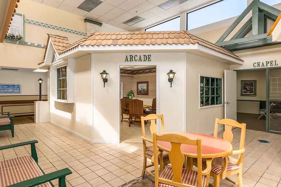 Interior common area of an assisted living facility featuring a mock storefront labeled 'ARCADE', a round table with chairs, benches, and a nearby chapel entrance.