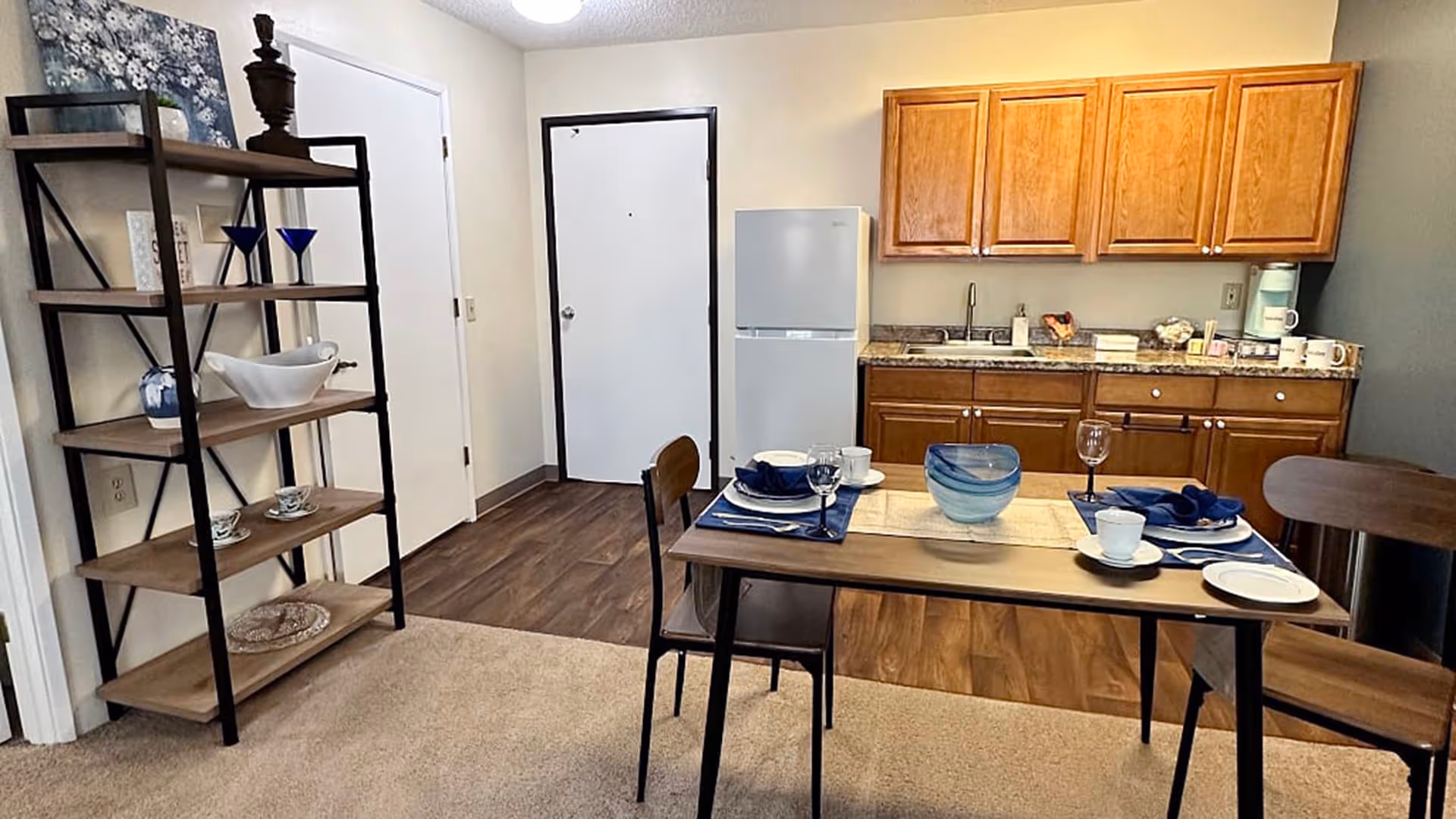 Interior view of a small kitchen and dining area in a senior living facility. The kitchen has wooden cabinets, a countertop with a sink, a white refrigerator, and various kitchen items. In front of the kitchen is a dining table set for two with plates, cups, napkins, and a decorative bowl. To the left, there is a black metal and wood shelving unit with decorative items and dishes. The floor transitions from carpet in the dining area to wood flooring in the kitchen and entryway.