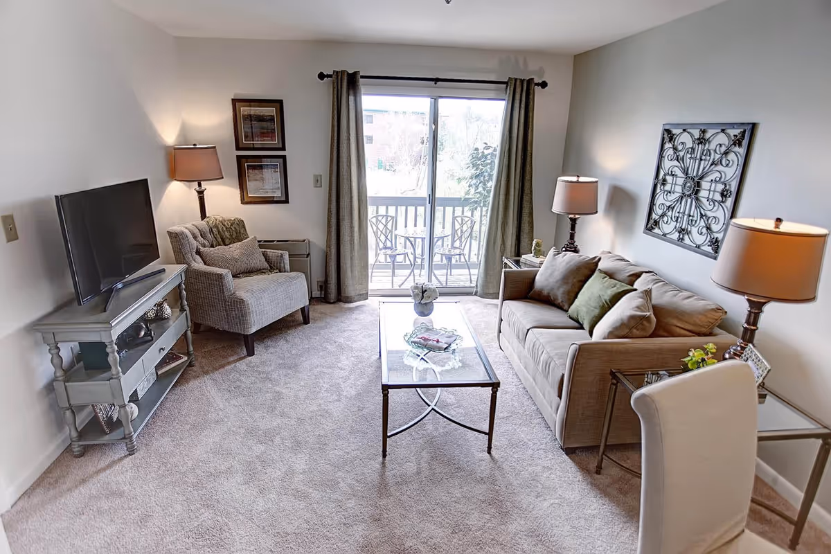 A cozy living room with a beige sofa adorned with cushions, a glass coffee table with a decorative item, a cushioned armchair, a TV on a stand, two table lamps, and a sliding glass door leading to a balcony with two chairs and a small table outside.