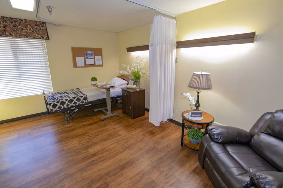 A well-lit room with a hospital bed covered with a patterned blanket, a small wooden nightstand with a vase of white flowers, a bulletin board on the wall, a window with blinds and a floral valance, a white privacy curtain, a round side table with a lamp and a small plant, and a dark brown leather recliner chair.