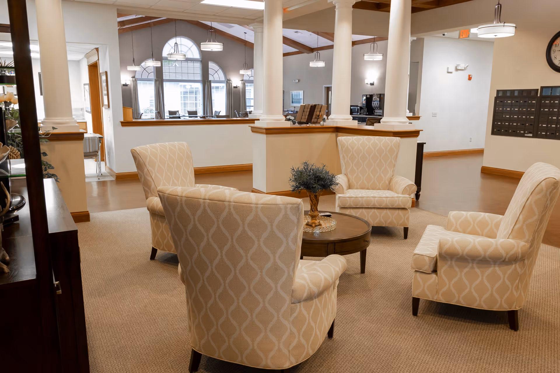 A cozy seating area in a senior living facility with four beige patterned armchairs arranged around a round wooden coffee table with a small plant centerpiece. The space features beige carpeting, white columns, and large windows with arched tops letting in natural light. The background shows a dining area with tables and chairs, pendant lights, and a clock on the wall.