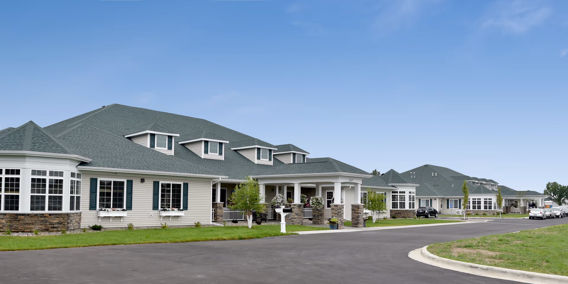 Exterior view of Birchwood Cottages - Owatonna, showing a large single-story building with multiple windows, green roof, and a covered entrance with stone pillars. Several cars are parked along the driveway and the sky is clear and blue.
