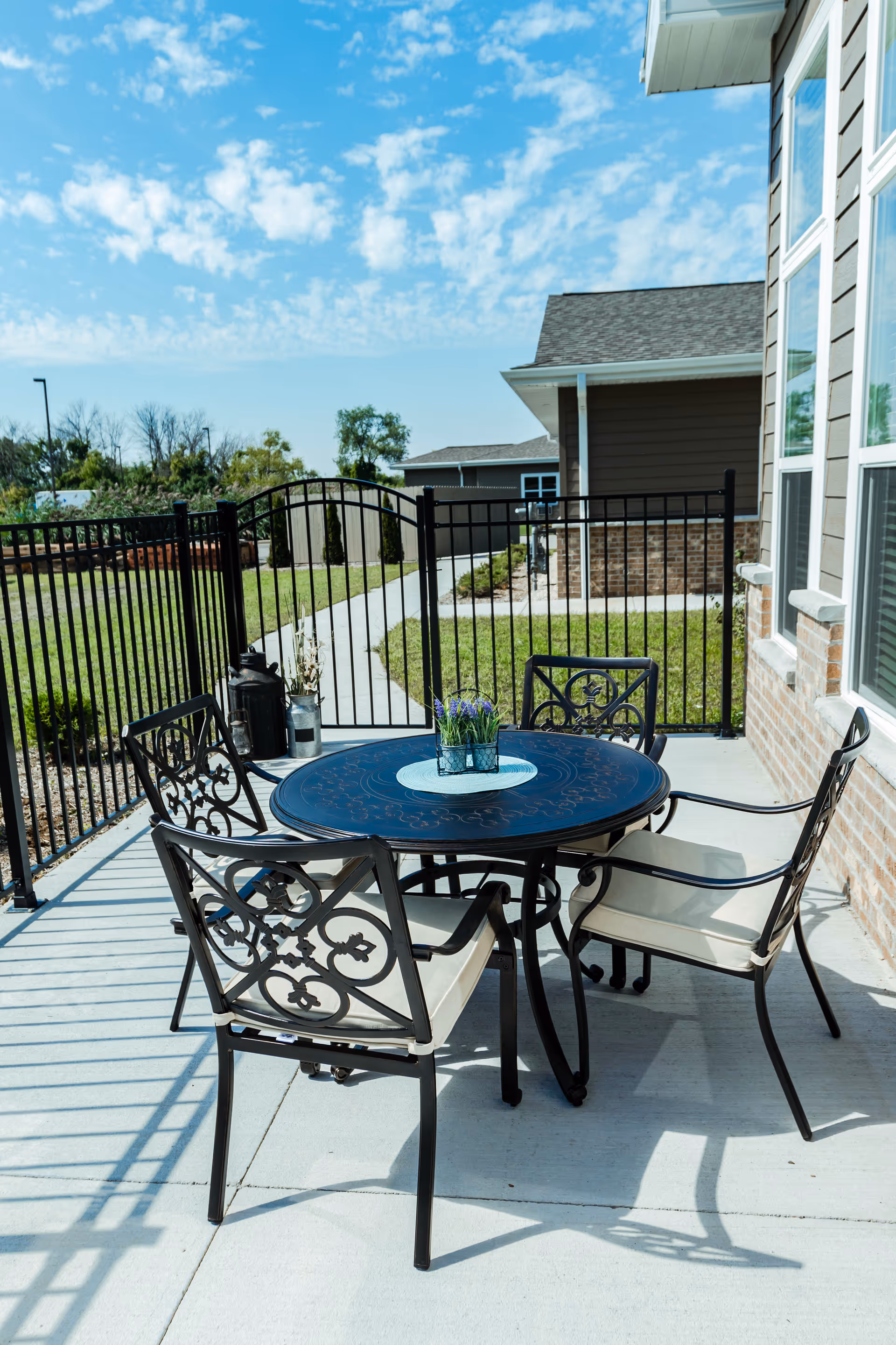 Outdoor patio area with a round black metal table and four matching chairs with beige cushions. The patio is enclosed by a black metal fence, and there is a small potted plant on the table. The background shows a walkway, green grass, and parts of a building under a partly cloudy blue sky.