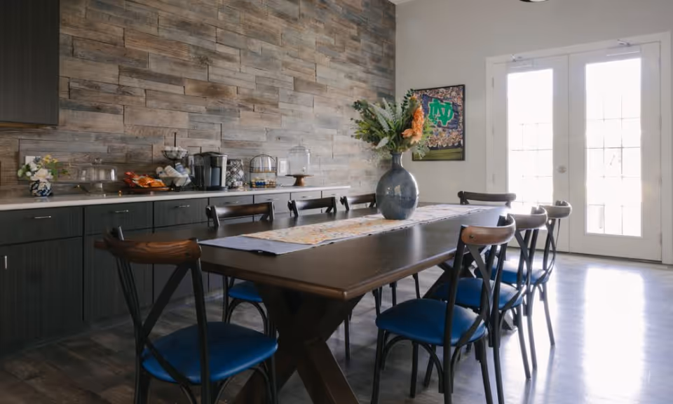 A bright communal dining room with a long wooden table surrounded by blue-seated chairs, a vase of flowers centerpiece, a wood-paneled accent wall and a countertop with snacks and appliances near French doors.