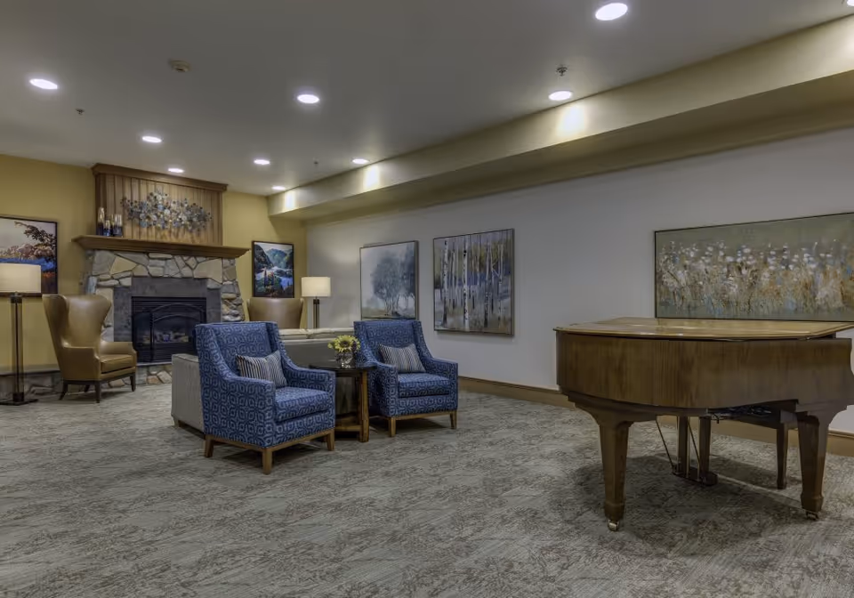 A cozy living room area with a stone fireplace, two blue patterned armchairs, a small wooden table with a flower arrangement, a grand piano, and several framed paintings on the walls. The room is softly lit with recessed ceiling lights and floor lamps.