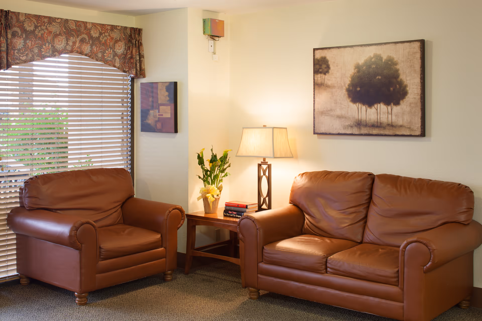 A cozy sitting area with a brown leather armchair and a matching two-seater sofa. Between them is a wooden side table with a lamp, a small plant, and a couple of books. The wall behind features a painting of trees, and there is a window with blinds and a patterned valance on the left side.