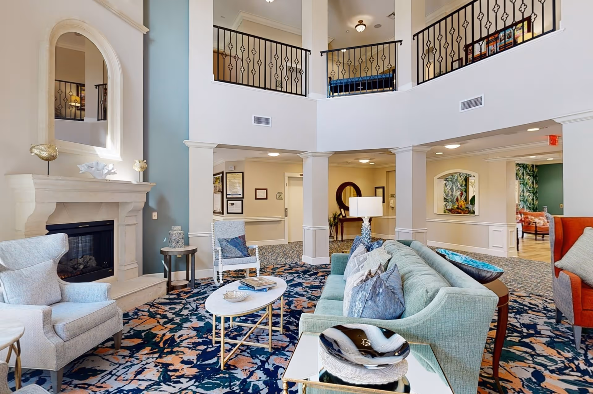A spacious and well-lit living room area in a senior living facility featuring a light blue sofa with multiple cushions, a patterned carpet, a white armchair, a small round coffee table, and a fireplace with a large mirror above it. The room has high ceilings with a second-floor balcony railing visible, and neutral-colored walls with some blue accent walls.