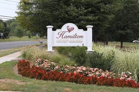 A white curved sign with Hamilton lettering, surrounded by colorful flowers outdoors.