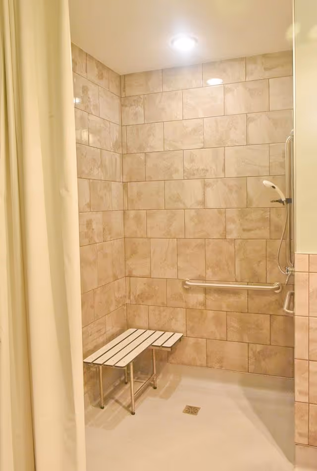 A tiled shower area with beige tiles on the walls, a white shower bench with metal legs, a handheld showerhead mounted on the wall, a grab bar, and a ceiling light. A white shower curtain is partially visible on the left side.