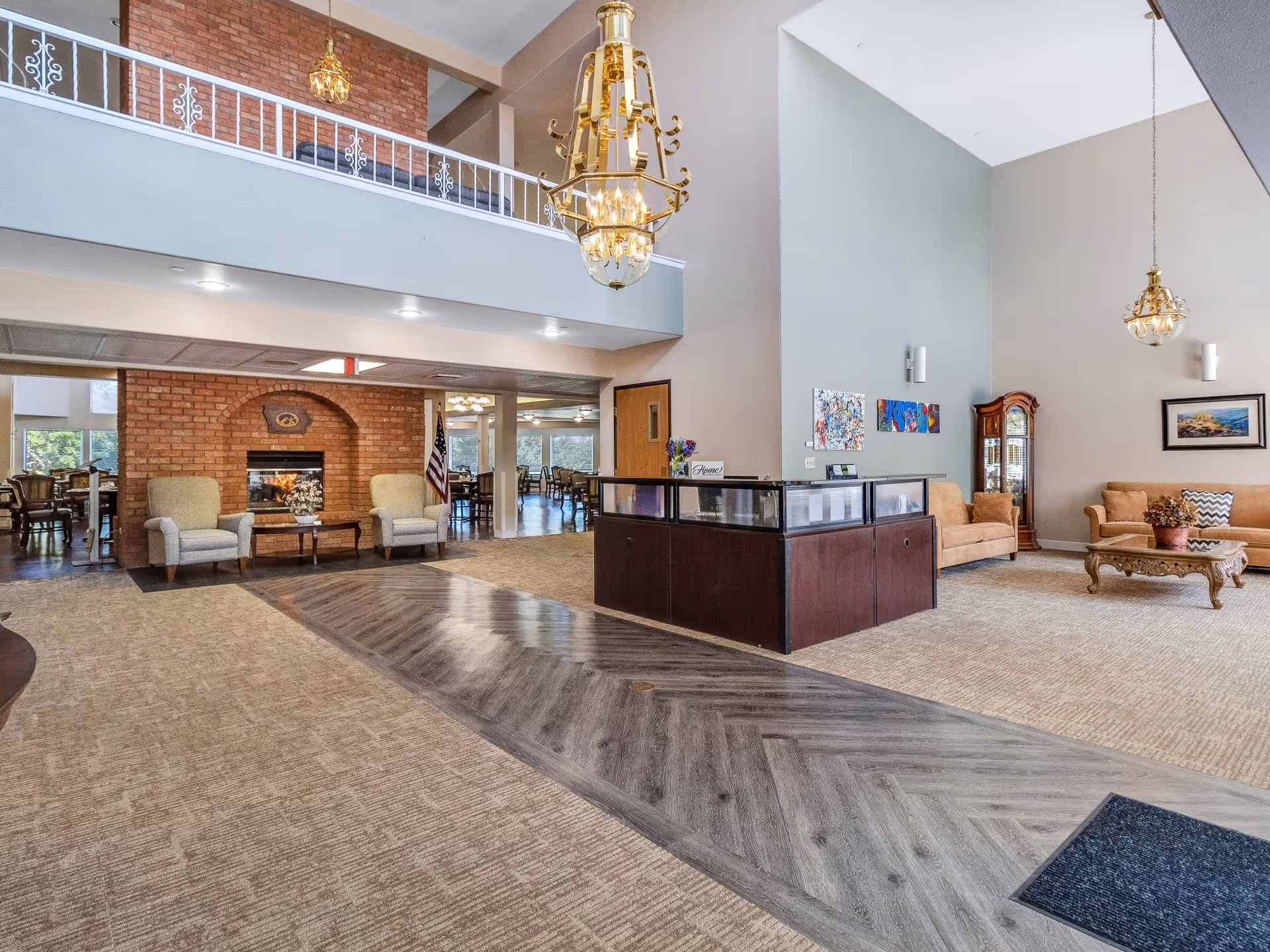 Spacious senior living facility lobby with high ceilings, a chandelier, seating areas including armchairs and sofas, a wooden reception desk, a brick fireplace, and decorative artwork on the walls.