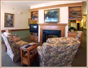 A cozy living room area with floral upholstered armchairs arranged around a wooden coffee table. There is a wooden fireplace with a framed painting above it, flanked by built-in wooden shelves and cabinets. A television is placed on a stand to the left of the fireplace. The walls are decorated with framed artwork, and the room has a warm, inviting atmosphere.