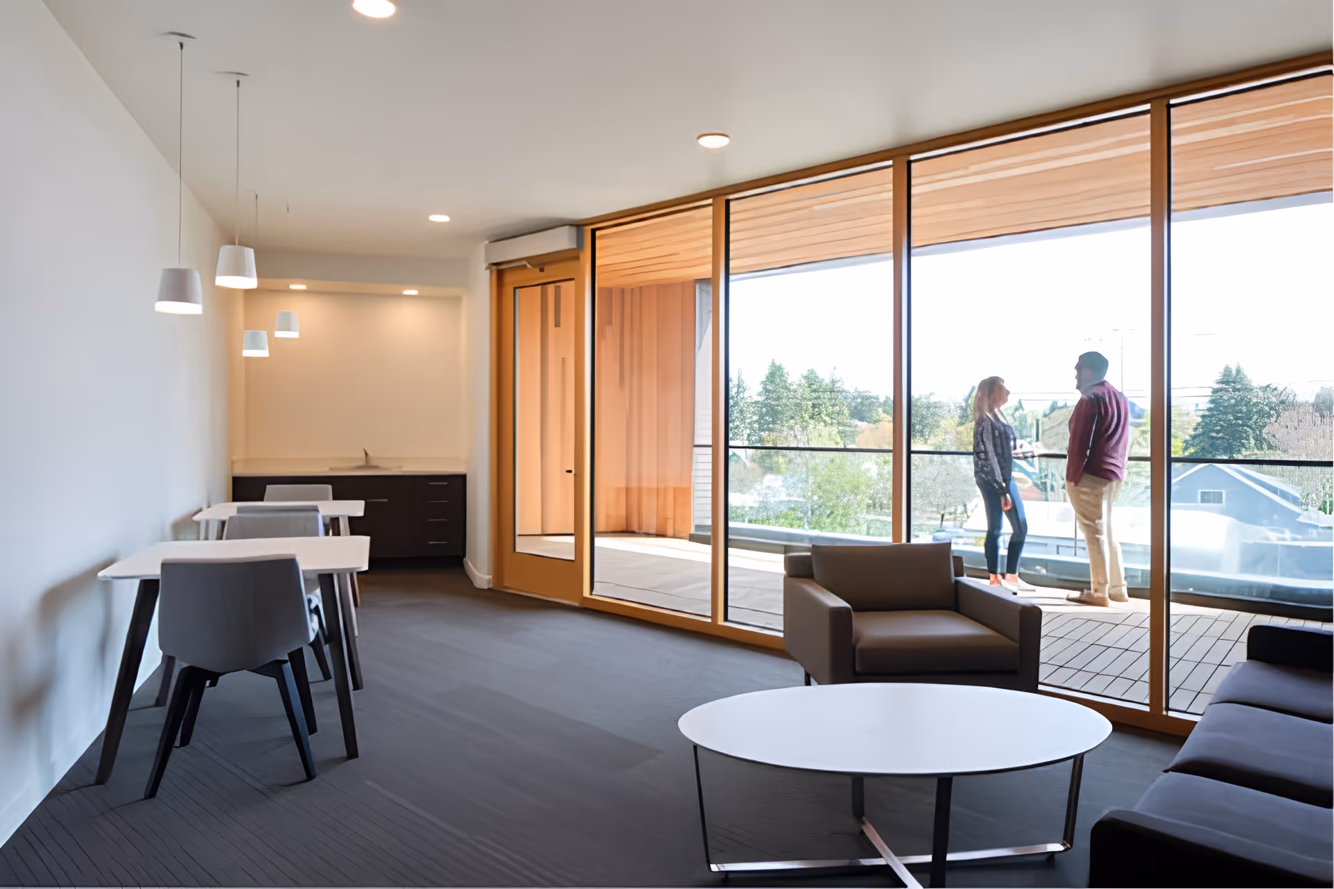 A modern common area with large floor-to-ceiling windows showing an outdoor balcony where two people are standing and talking. Inside, there are tables with chairs along the left wall, a small kitchenette area in the back, and comfortable seating including an armchair and a sofa near a round white coffee table.