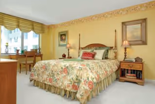 Sunlit bedroom with a floral bedspread on a wooden four-poster bed, matching nightstands, and a window seating/desk area.