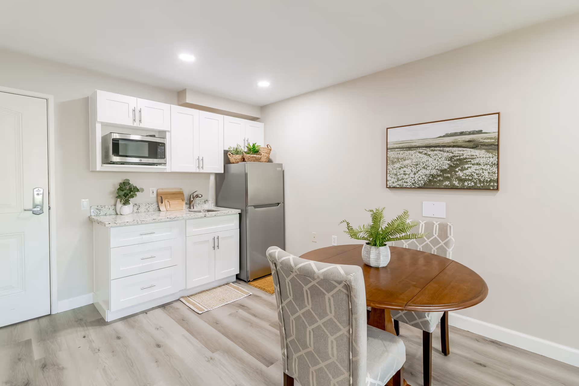 A small kitchen and dining area featuring white cabinets, a stainless steel refrigerator, a microwave, a sink, and a wooden dining table with two upholstered chairs. A potted plant is on the table, and a landscape painting hangs on the wall.